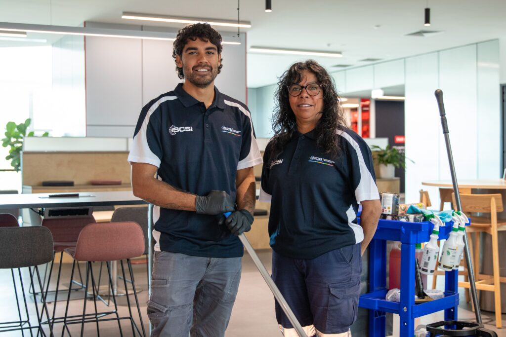 Two SCS Indigenous cleaning staff members smiling in a modern office environment, both wearing navy and white SCS Indigenous branded polo shirts. The male team member holds a mop and wears black gloves, while the female team member stands beside a blue cleaning trolley stocked with spray bottles and supplies.