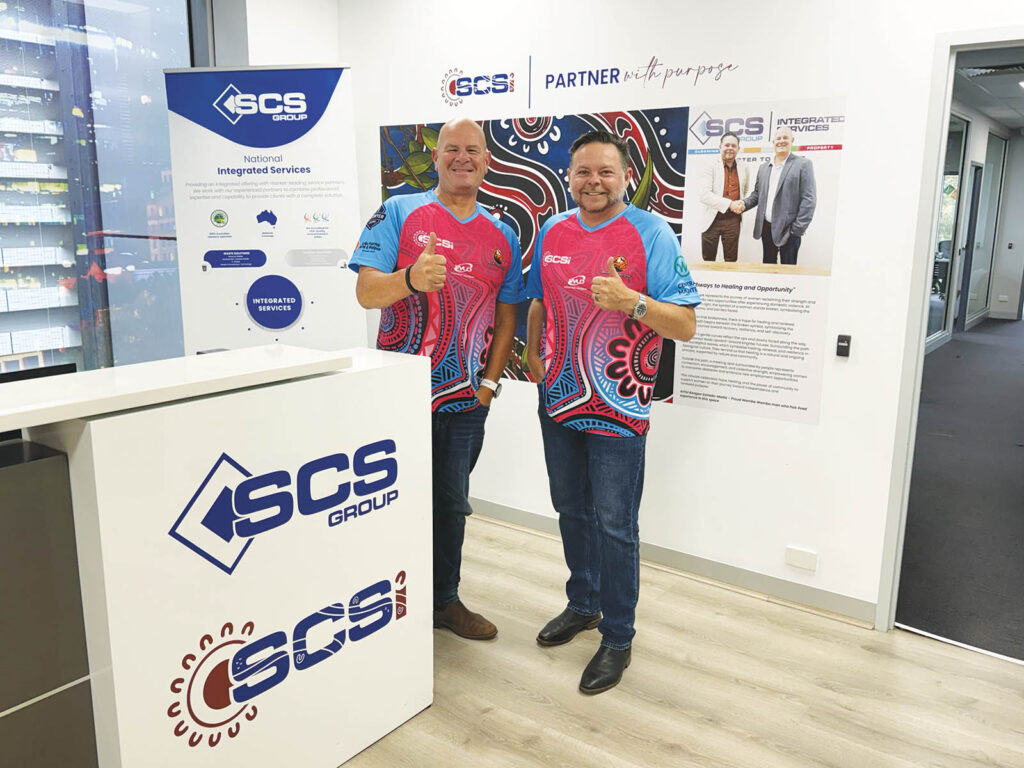 SCS Group Co-Founders Nicholas Pastras and Scott McCartney giving thumbs up in matching pink, blue, and black Aboriginal art-inspired jerseys inside the SCS Group office, with the company reception desk in the foreground displaying the SCS Group and SCS Indigenous logos, branded pull-up banners, and a "Partner with Purpose" wall display featuring Aboriginal artwork in the background.